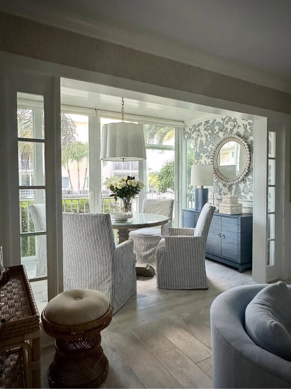 Coastal dining room with a round wood table, striped slipcovered chairs, blue sideboard, and floral wallpaper