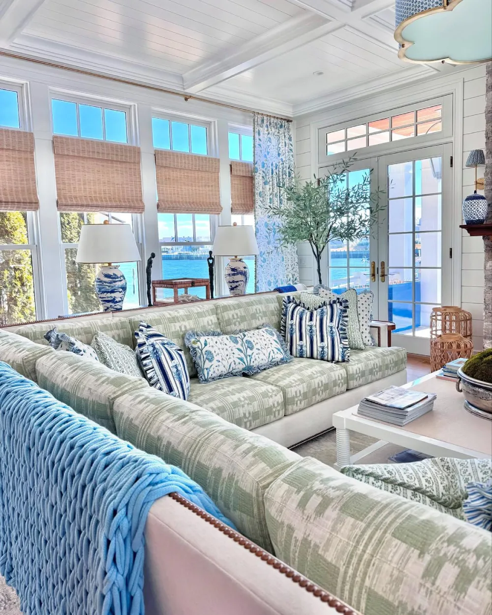 Coastal living room with pale blue sofa, woven shades, patterned pillows, and water views