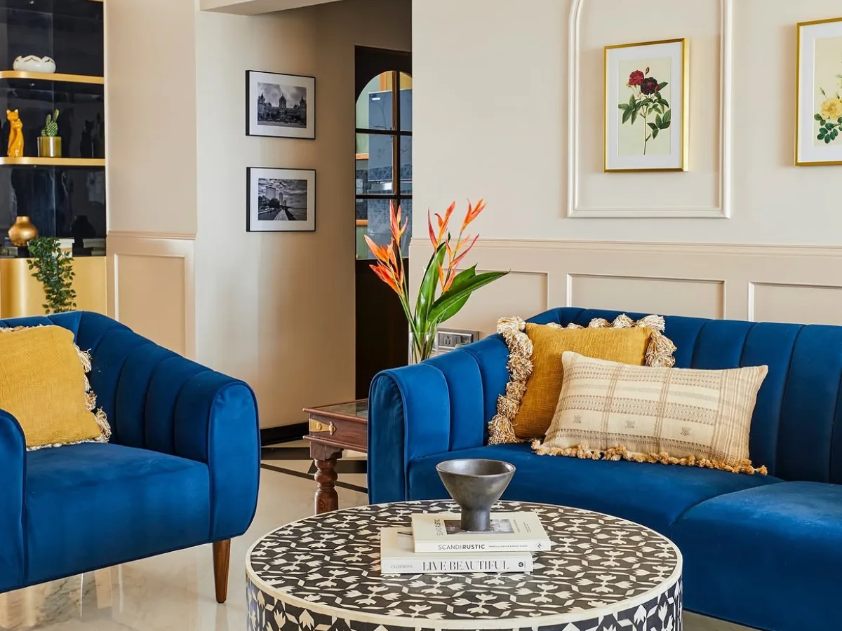 Elegant living room with blue velvet sofas, framed botanical artwork, decorative wall molding, and a patterned round coffee table.