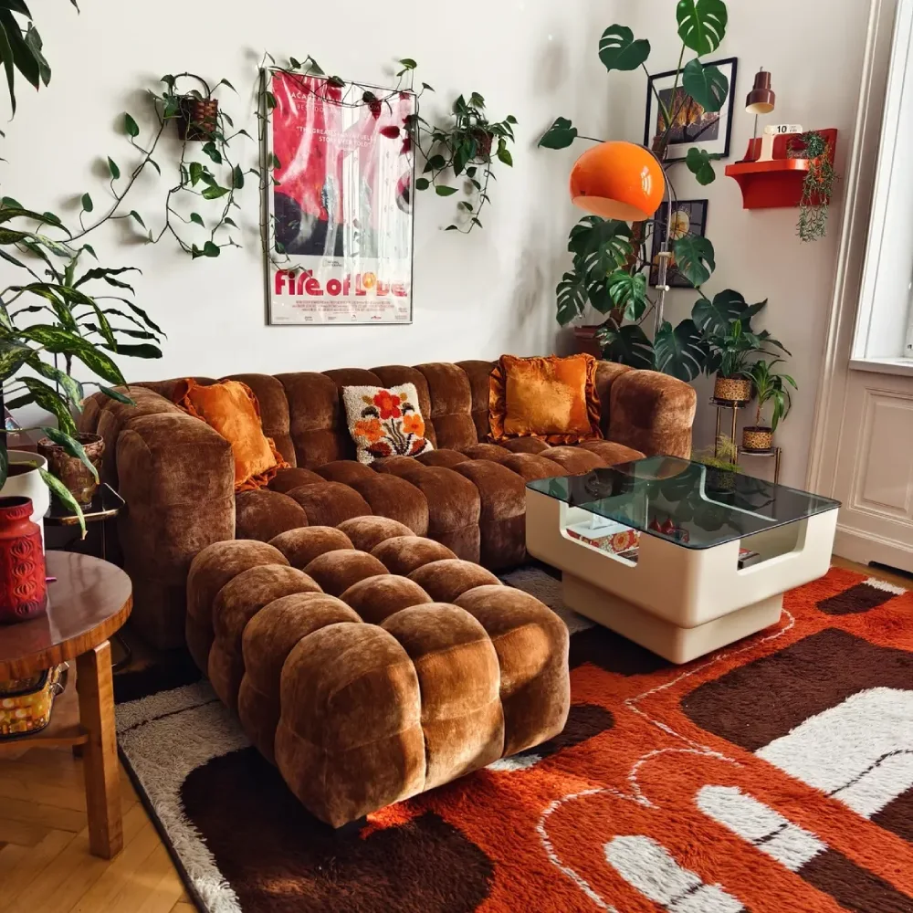 Brown velvet sofa with orange pillows, tropical plants, and a colorful patterned rug