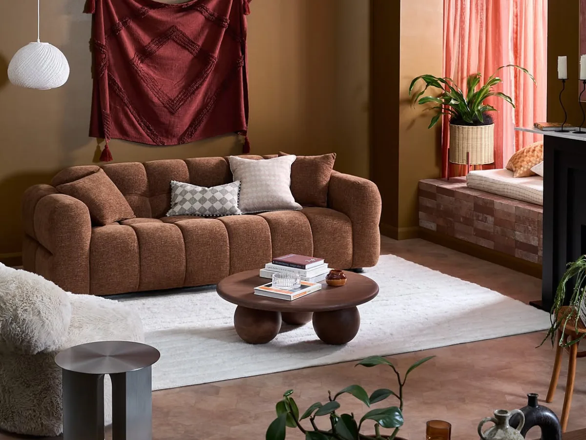 Warm living room with a brown sofa, burgundy wall hanging, white rug, wood coffee table, plants, and coral curtains