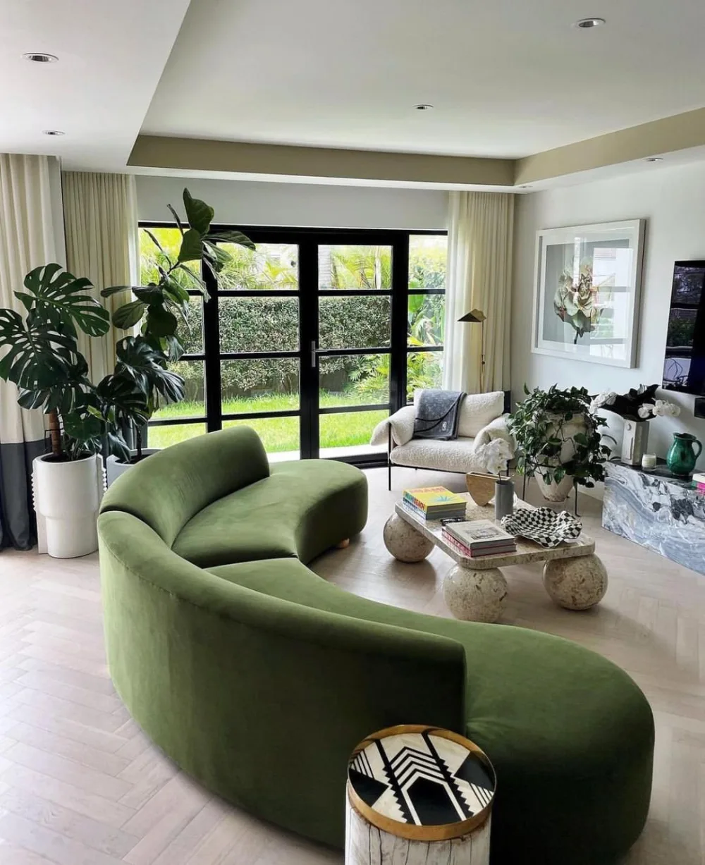 Curved sage green sofa in a bright living room with large windows and indoor plants