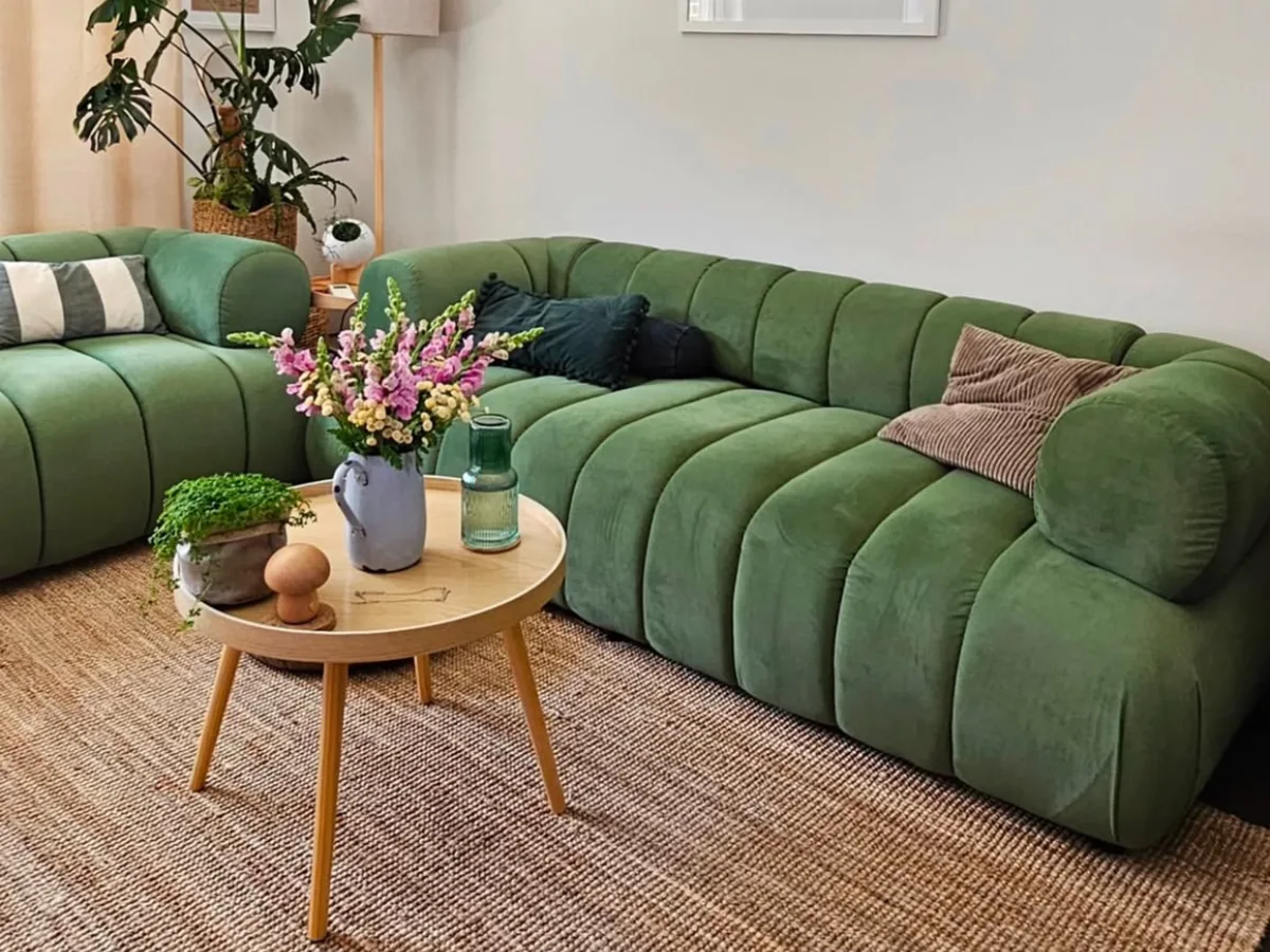 Green curved sectional sofa with striped pillows, a round wooden coffee table, plants, and floral decor in a cozy living room.
