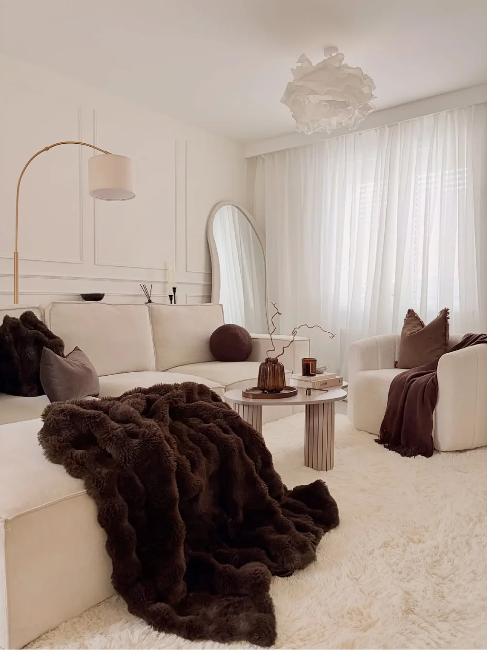 cozy cream living room with dark brown throws, plush rug, curved mirror, and warm lighting