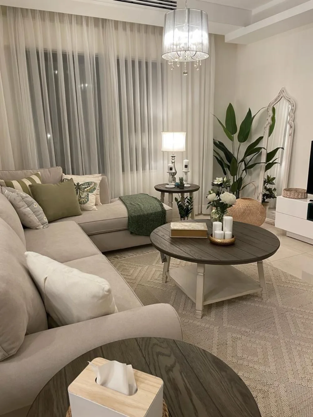neutral living room with sage green pillows, plants, dark wood coffee table, and soft lighting