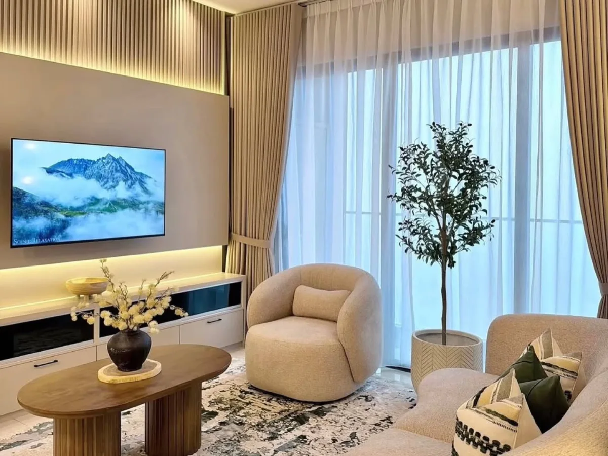 Cozy modern living room with beige seating, wood slat walls, sheer curtains, warm lighting, and a large indoor tree.