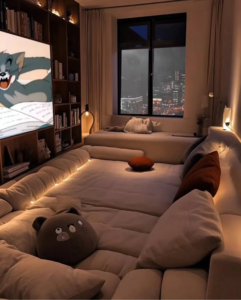 Large cream sectional sofa with pillows, warm ambient lighting, bookshelves, and city night view