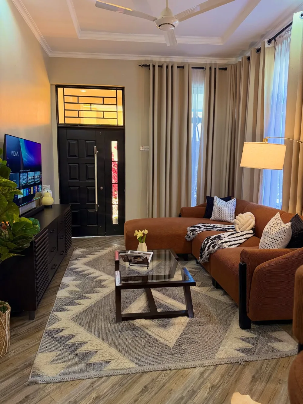 Cozy living room with brown seating, cream curtains, patterned rug, and layered pillows