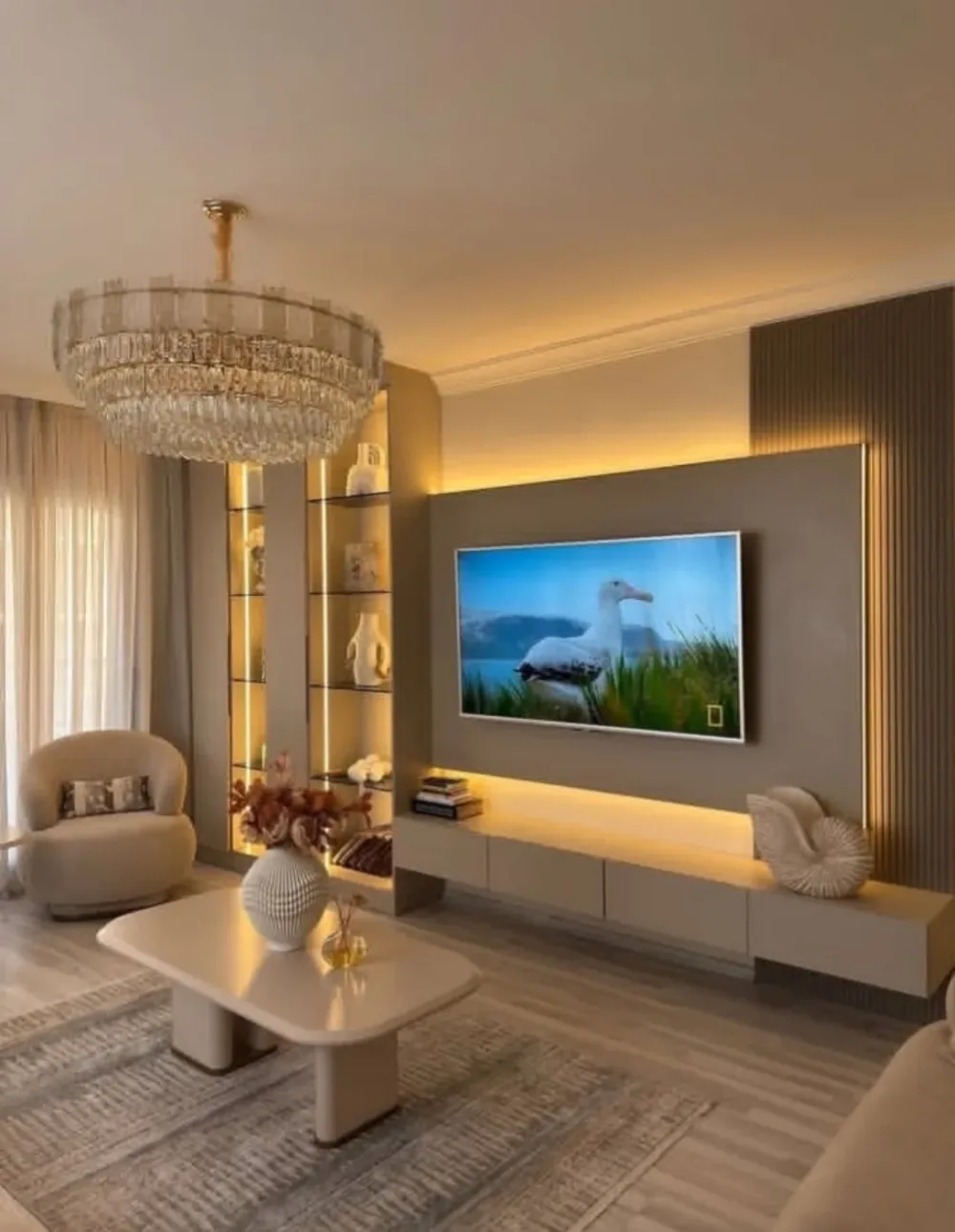 Elegant living room with warm ambient lighting, a crystal chandelier, built-in TV wall, and neutral furniture