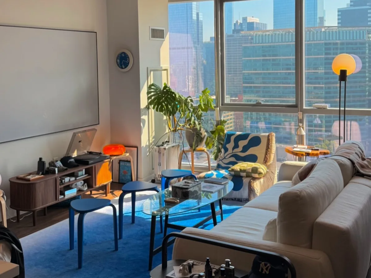 Bright apartment living room with a cream sofa, blue rug, patterned accent chair, plants, and floor-to-ceiling city views.