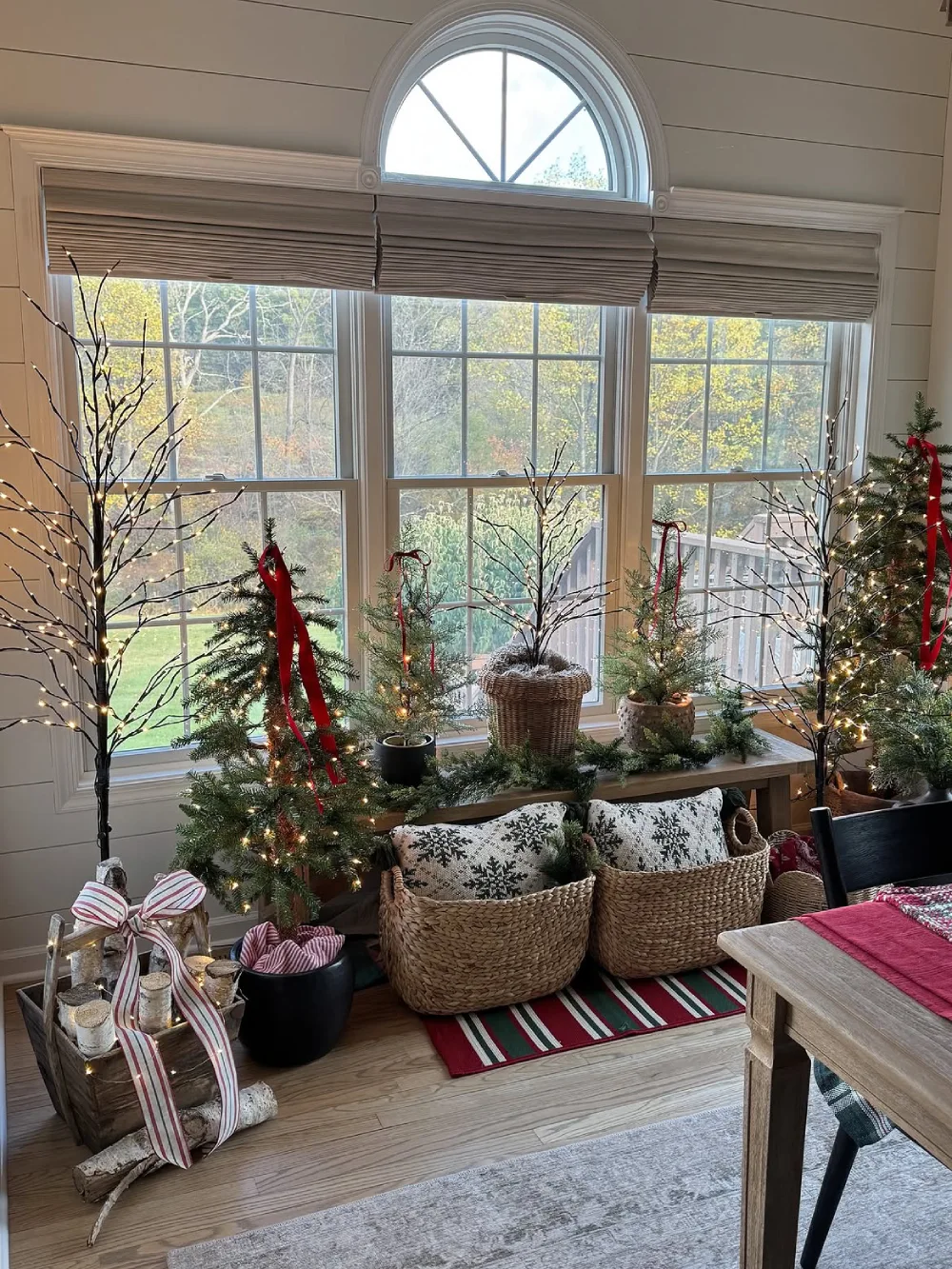 Bright winter living space with small Christmas trees, woven baskets, greenery, and red ribbons by large windows