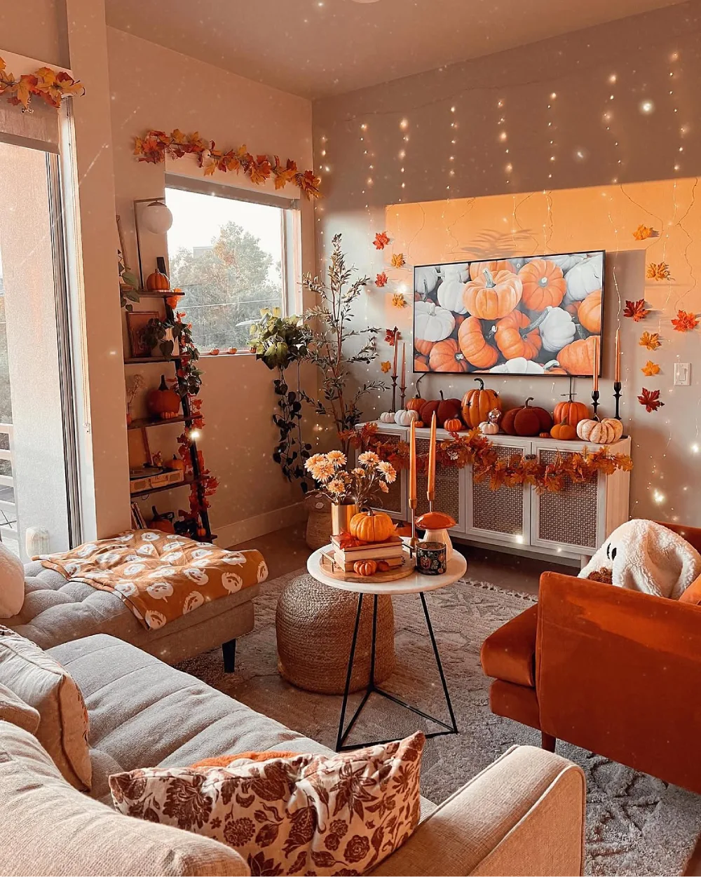 Fall living room decorated with orange and white pumpkins, autumn leaf garlands, warm fairy lights, and cozy neutral seating