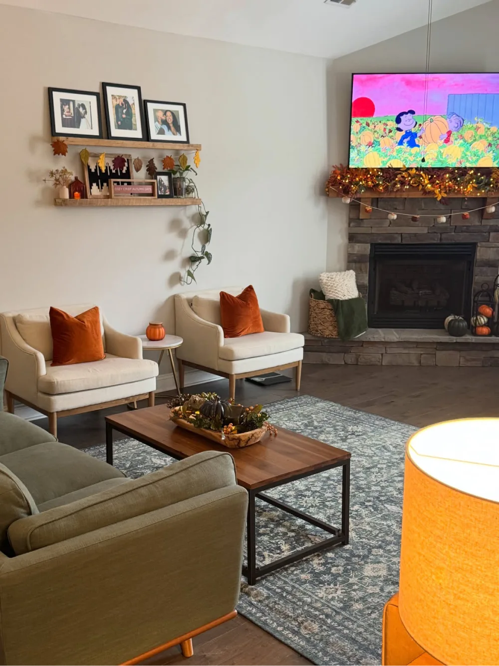 Simple fall living room with cream accent chairs, burnt-orange pillows, autumn garlands on floating shelves, and a stone fireplace decorated with seasonal pumpkins