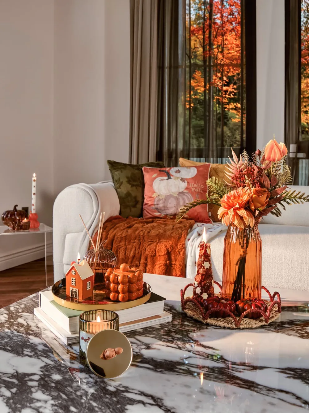 Cozy cream living room with rust-colored knitted blankets, fall-themed pillows, an amber glass vase of autumn flowers, and pumpkin decorations