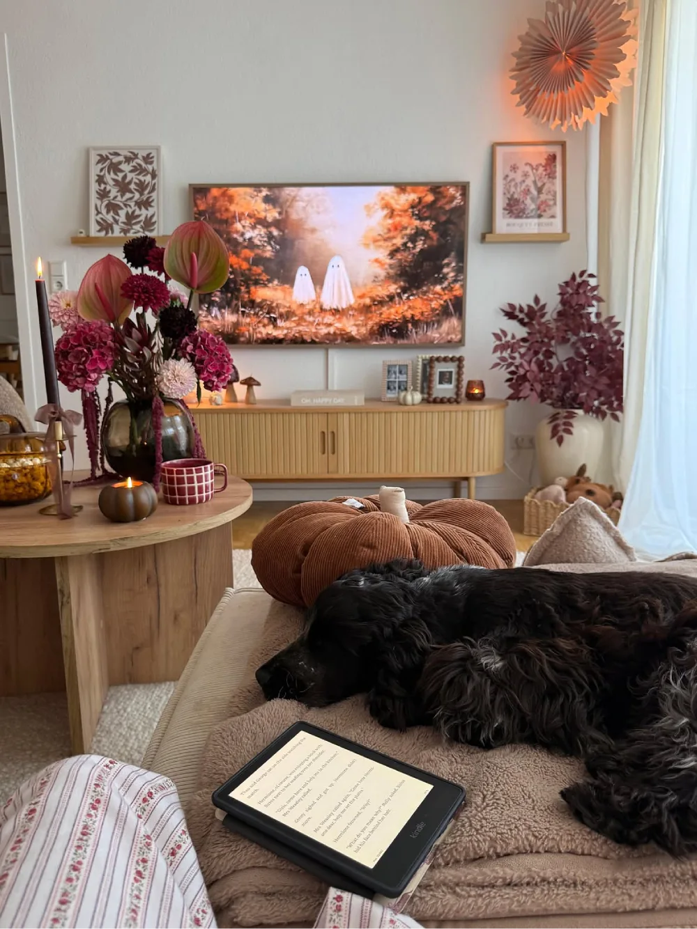 Cozy fall living room with a sleeping black dog on a beige sofa, a chunky brown pumpkin pillow, warm blankets, autumn flowers, and soft lighting