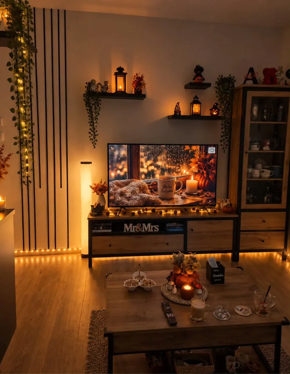 Dark and cozy living room with warm fairy lights, illuminated lanterns, autumn decorations on floating shelves, and a television displaying a rainy fall window scene