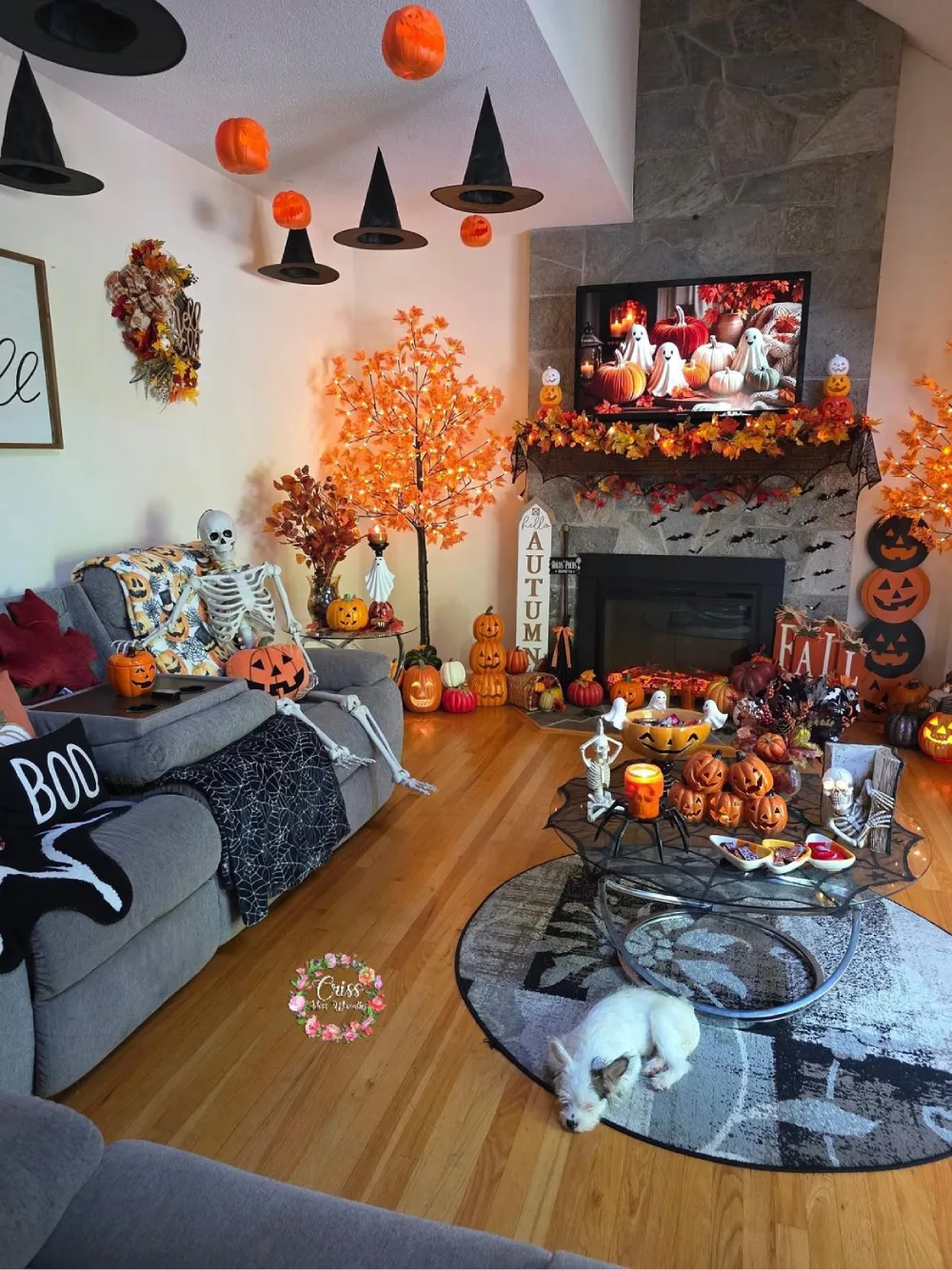 Halloween living room with a stone fireplace, hanging witch hats, orange pumpkin lanterns, skeleton decorations, and autumn leaf garlands
