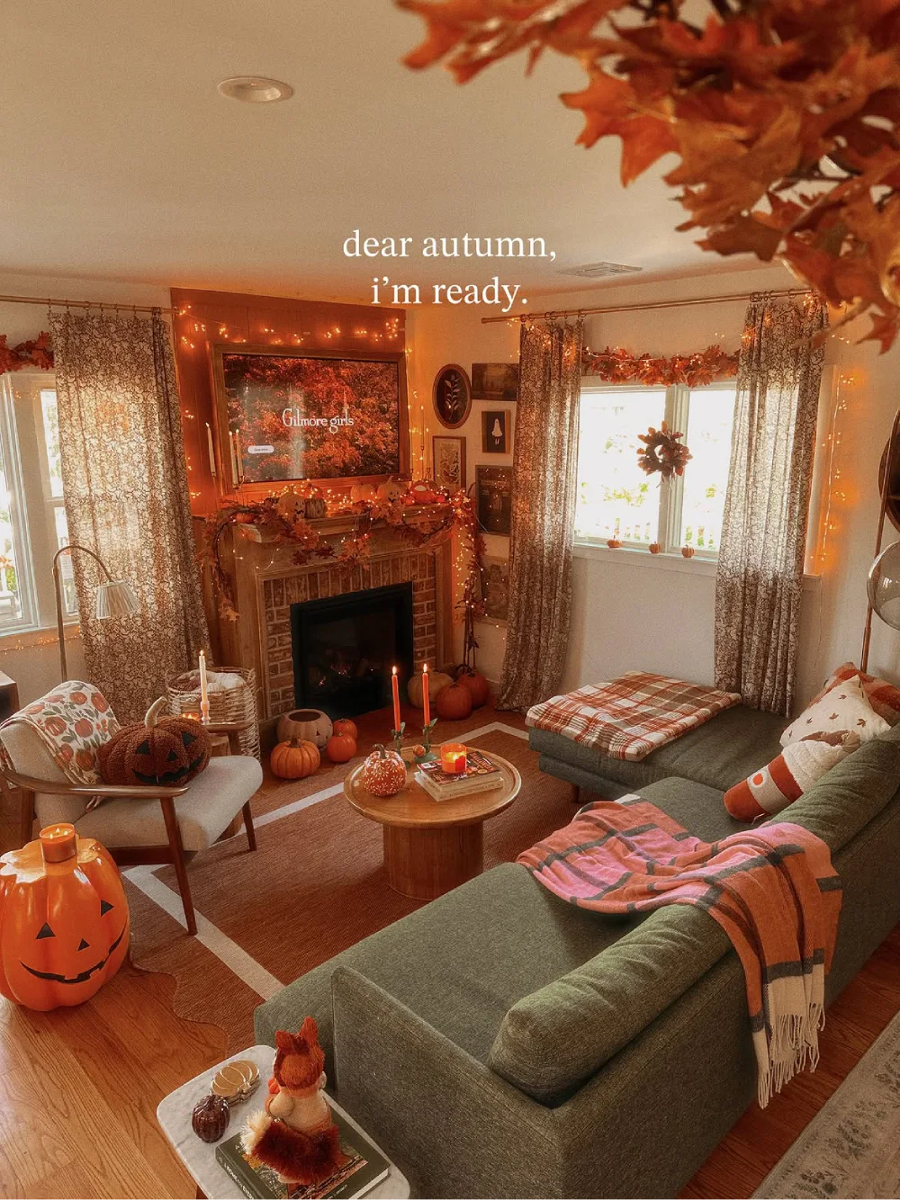 Autumn living room with a green sofa, pumpkin decorations, warm string lights, patterned curtains, and a fireplace surrounded by fall garlands