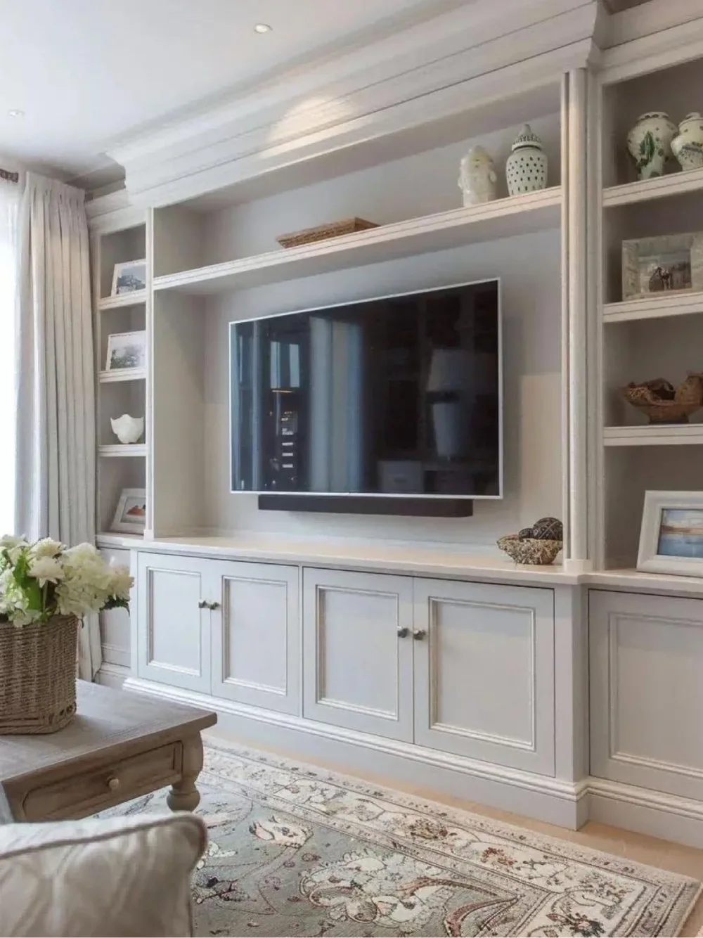 Elegant white built-in living room shelves surrounding a wall-mounted television, with decorative ceramics, framed photos, and closed storage cabinets.