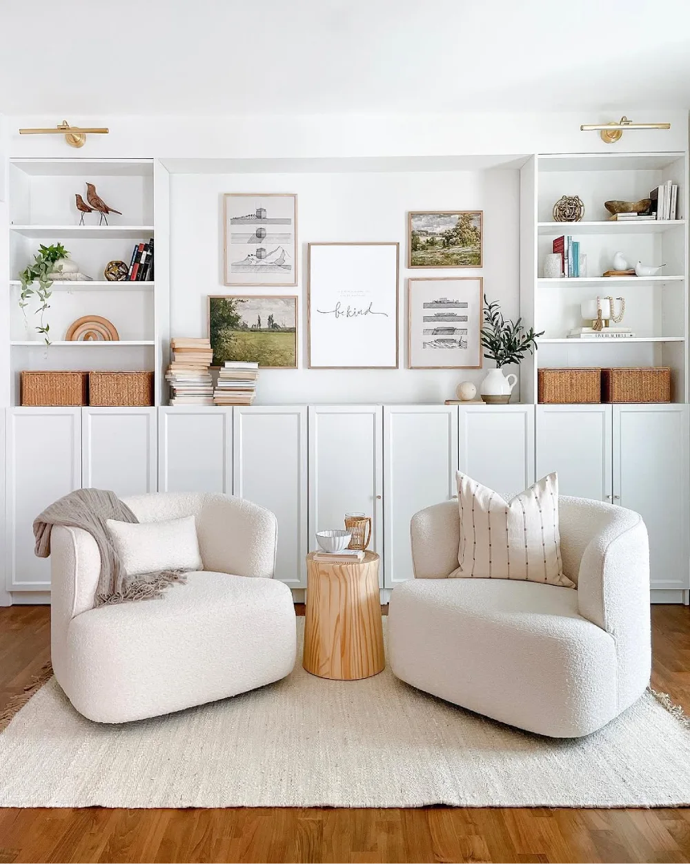 Minimalist white built-in living room shelves with closed lower cabinets, woven storage baskets, framed artwork, books, greenery, and two cream accent chairs.