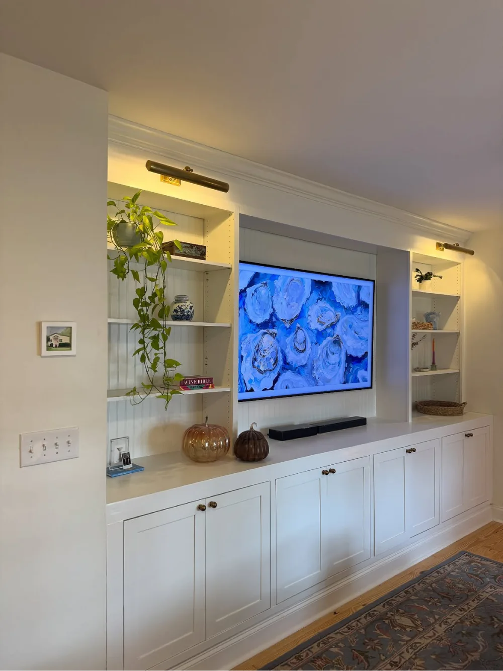 White built-in entertainment center with a wall-mounted television, open shelves displaying books and plants, brass picture lights, and spacious lower storage cabinets.