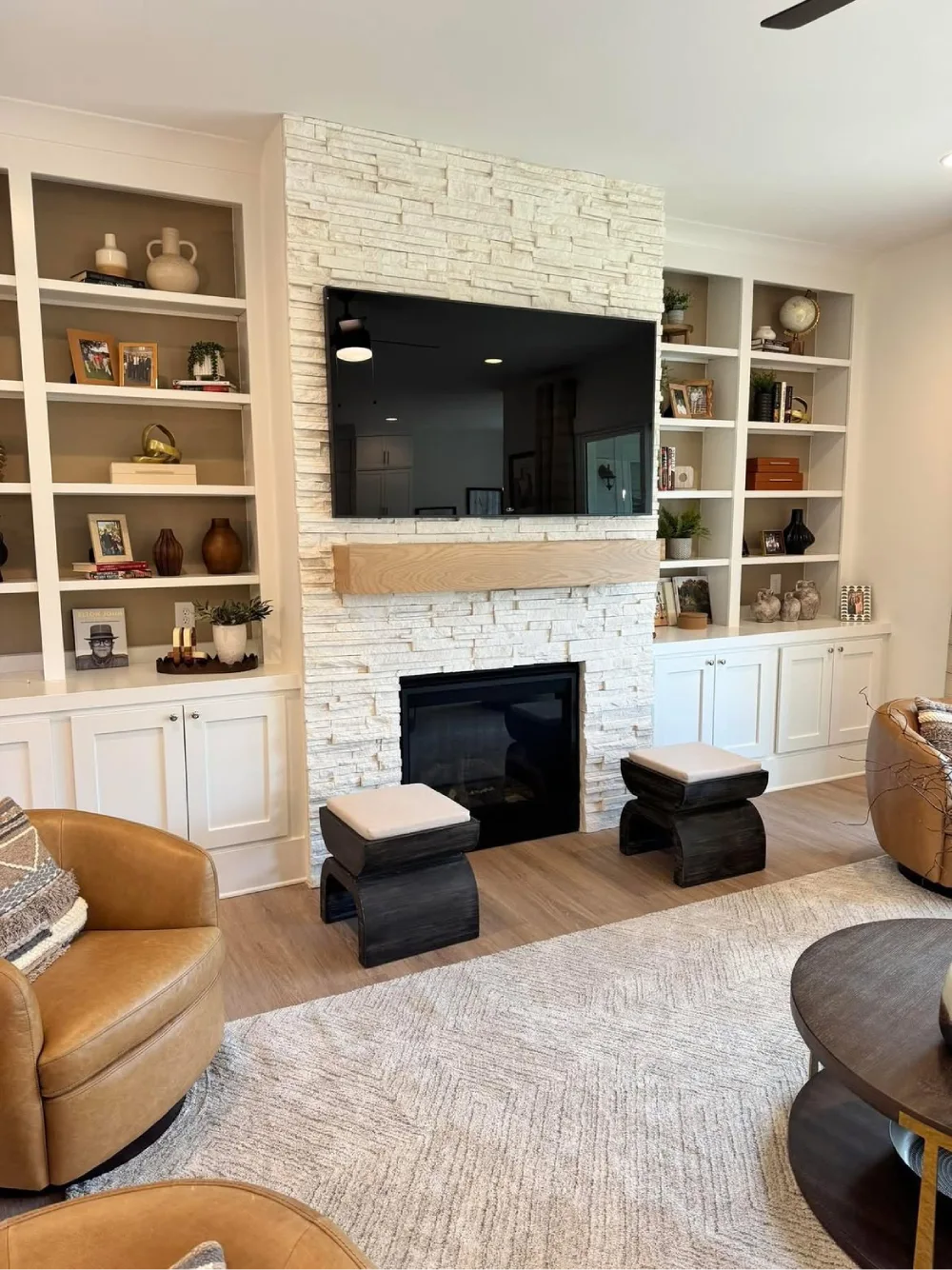 White built-in bookshelves framing a textured stone fireplace with a wall-mounted television, wooden mantel, decorative pottery, framed photos, and lower cabinets.