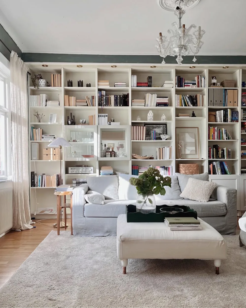 Wall-to-wall white built-in living room bookshelves filled with books, decorative objects, storage boxes, and framed artwork behind a comfortable neutral sofa.