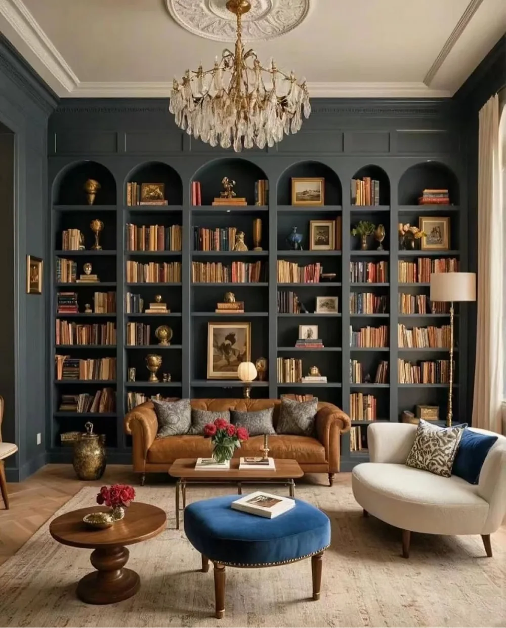 Dramatic charcoal-blue built-in bookcases with arched upper compartments, rows of books, antique-style decorations, and a brown leather sofa beneath a crystal chandelier.