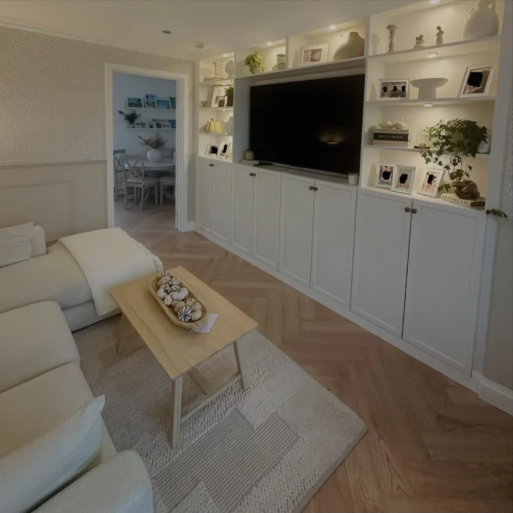 Modern white built-in living room shelves surrounding a large wall-mounted television, with decorative vases, framed photographs, small plants, and closed lower cabinets.