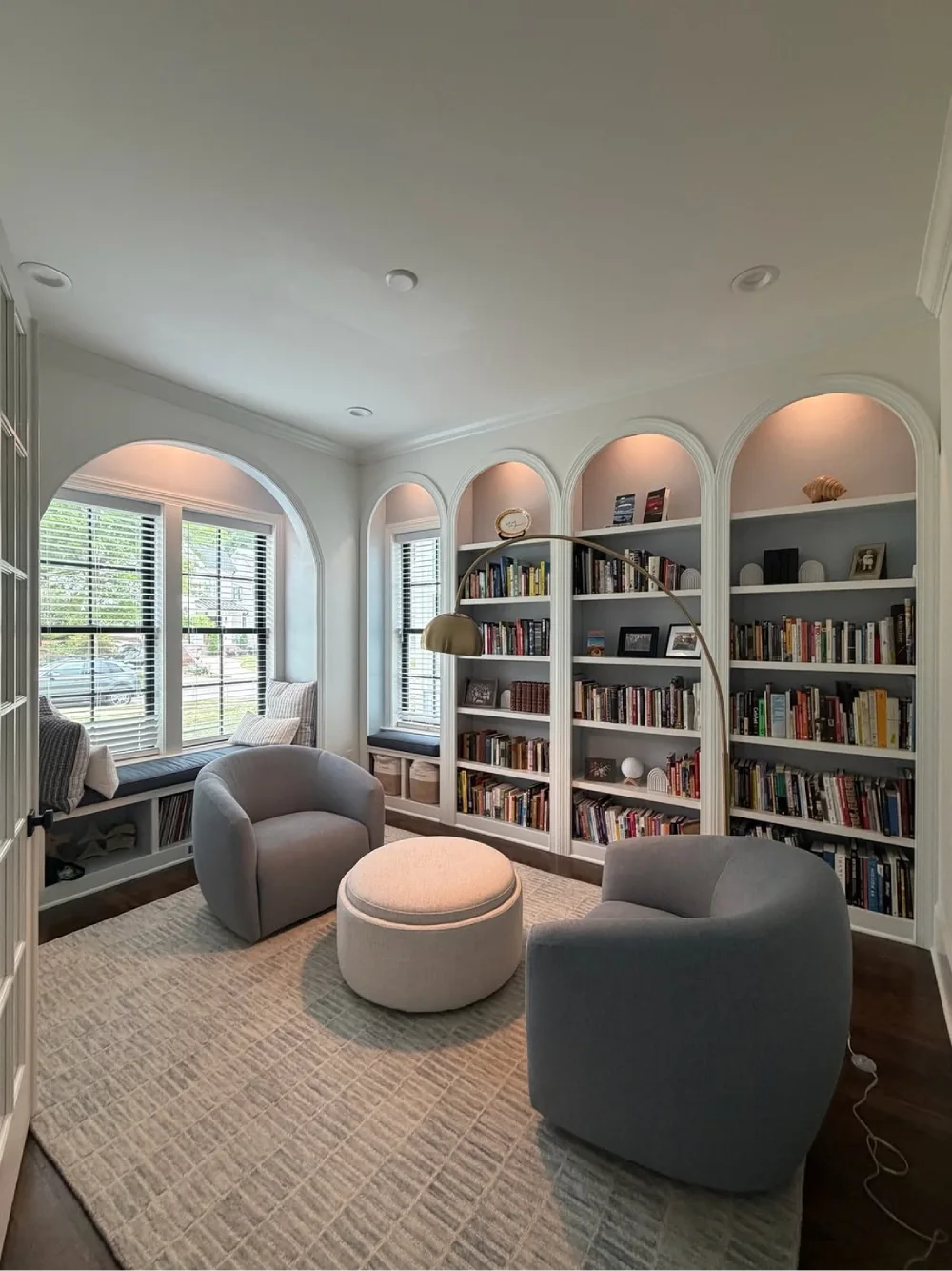 Cozy living room reading nook with gray armchairs, tall built-in bookshelves featuring arched niches, warm accent lighting, and a window seat with storage.