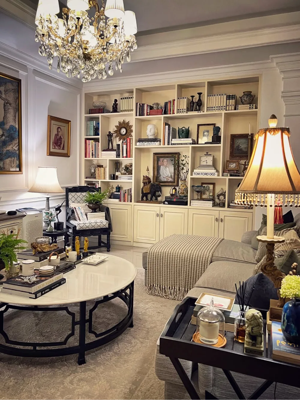 Traditional living room with cream built-in shelves and lower cabinets displaying books, framed portraits, sculptures, decorative ceramics, and collectibles beneath a crystal chandelier.