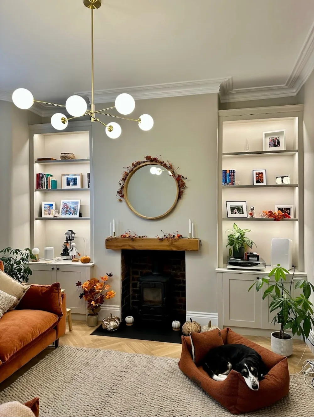 Living room fireplace framed by illuminated built-in shelves with books, family photos, potted plants, and closed cabinets, paired with a wooden mantel and warm autumn accents.