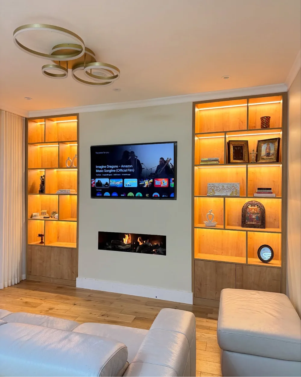 Modern living room with warm wood built-in display shelves, golden accent lighting, decorative artwork, books, and closed lower cabinets surrounding a television and linear fireplace.