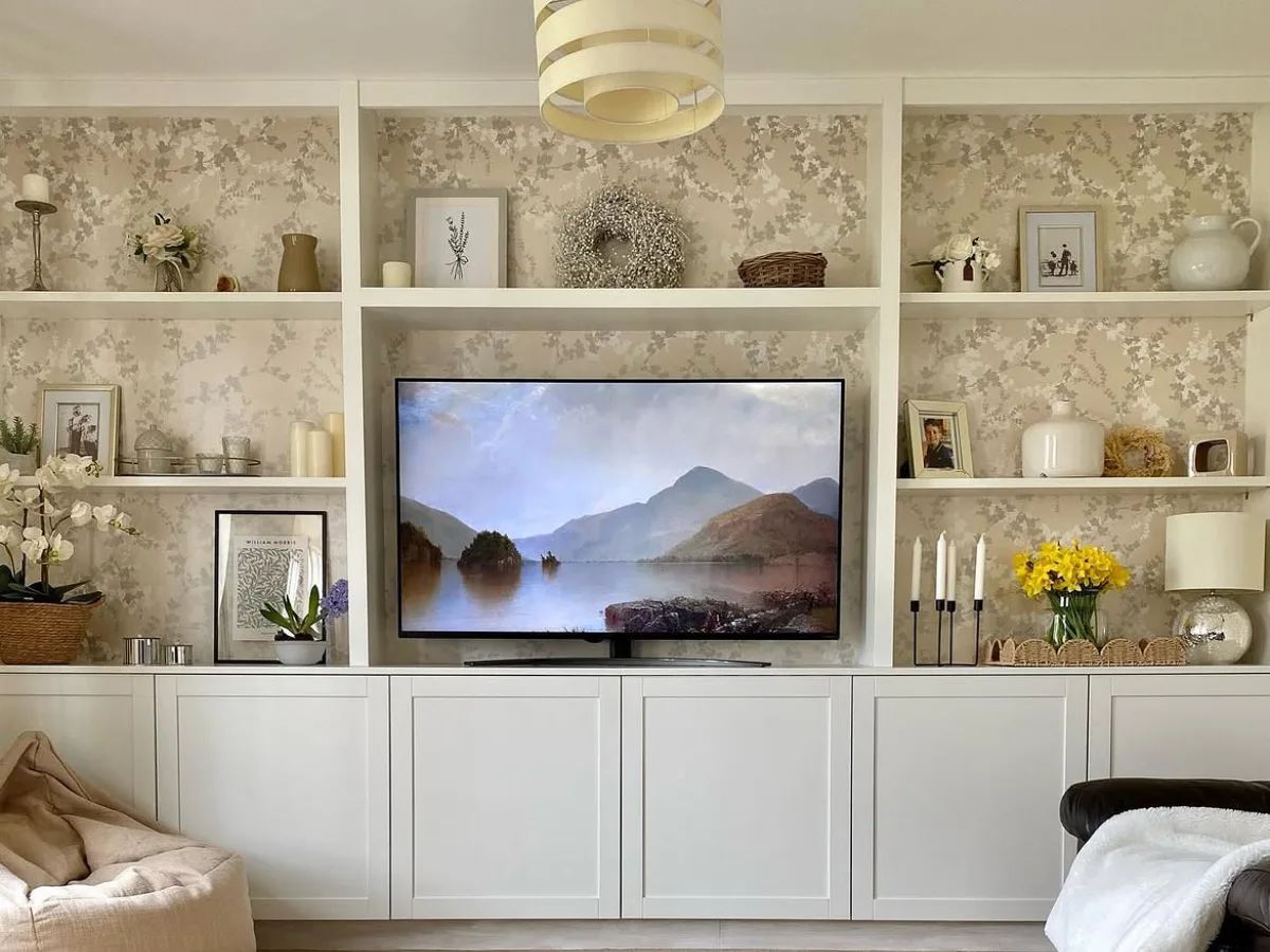 White built-in living room shelves surrounding a television, styled with framed artwork, decorative vases, flowers, and floral wallpaper above white cabinets.