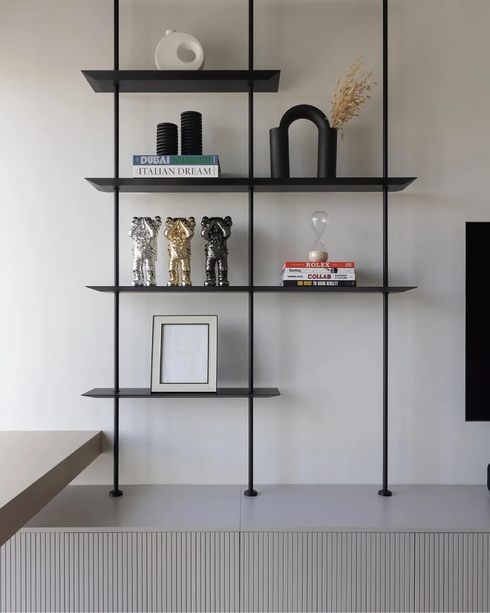 Modern black metal shelving system with slim horizontal shelves displaying sculptural vases, decorative figurines, books, and framed artwork above a white fluted cabinet