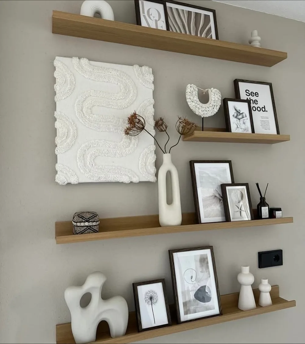 Long natural wood floating shelves arranged at different heights on a beige living room wall, displaying minimalist artwork, white ceramic vases, framed prints, and dried branches