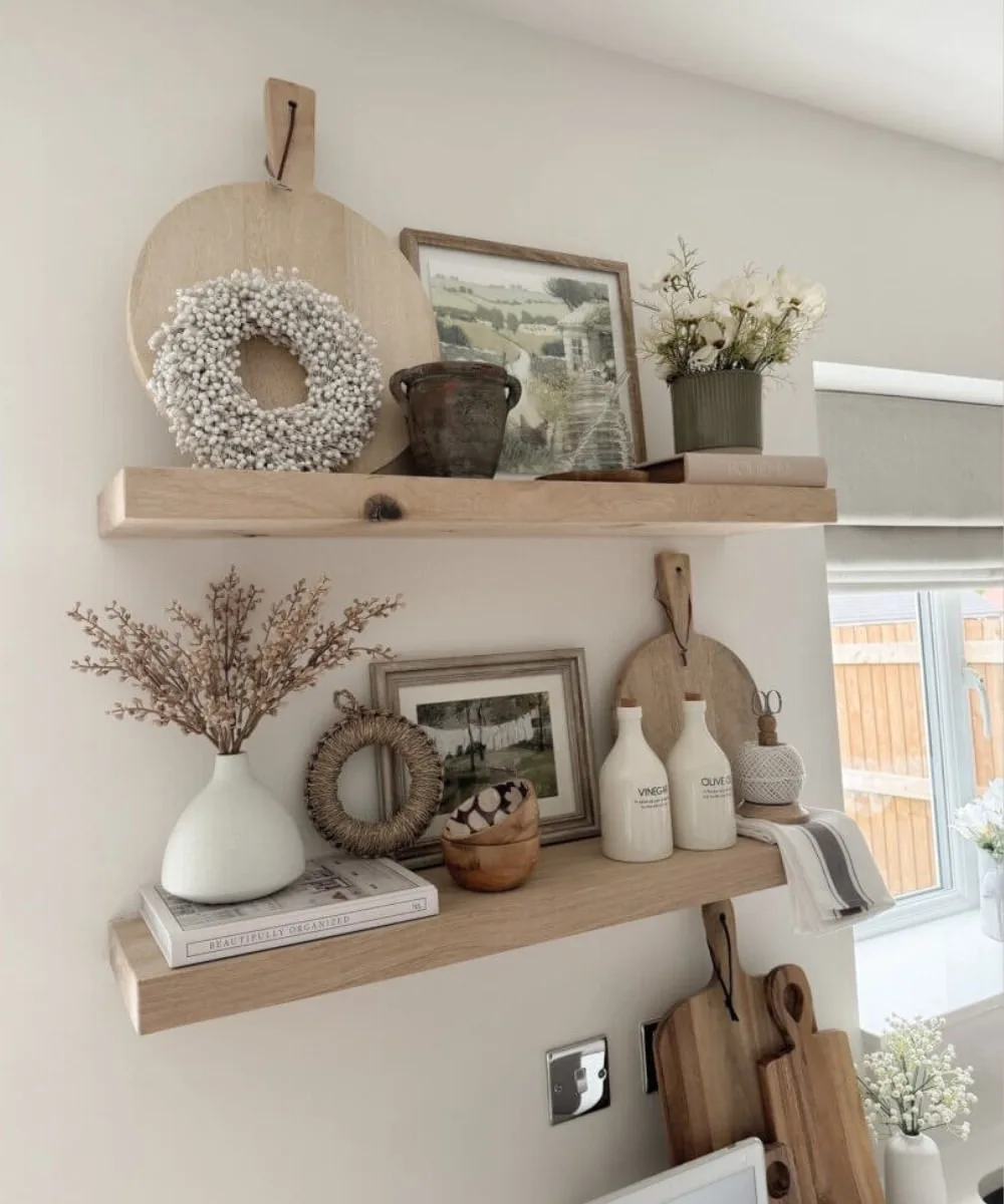 Rustic natural wood floating shelves styled with framed landscape prints, white ceramic vases, dried flowers, woven wreaths, wooden cutting boards, and neutral farmhouse decor