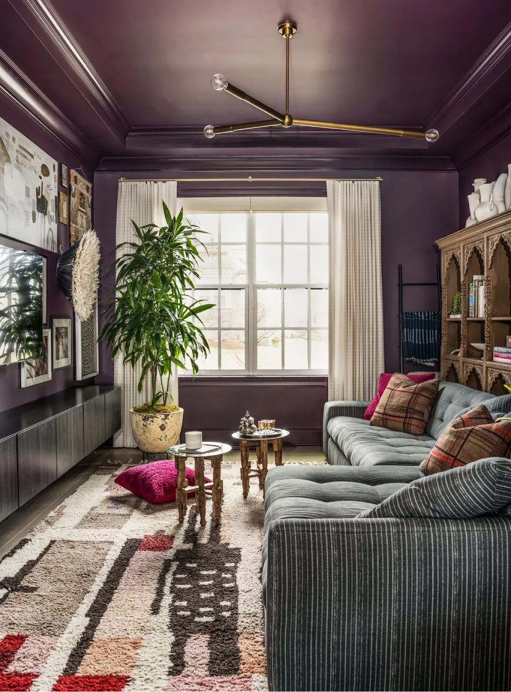 Moody living room with deep purple walls, a gray striped sectional sofa, colorful cushions, and an ornate wooden bookcase