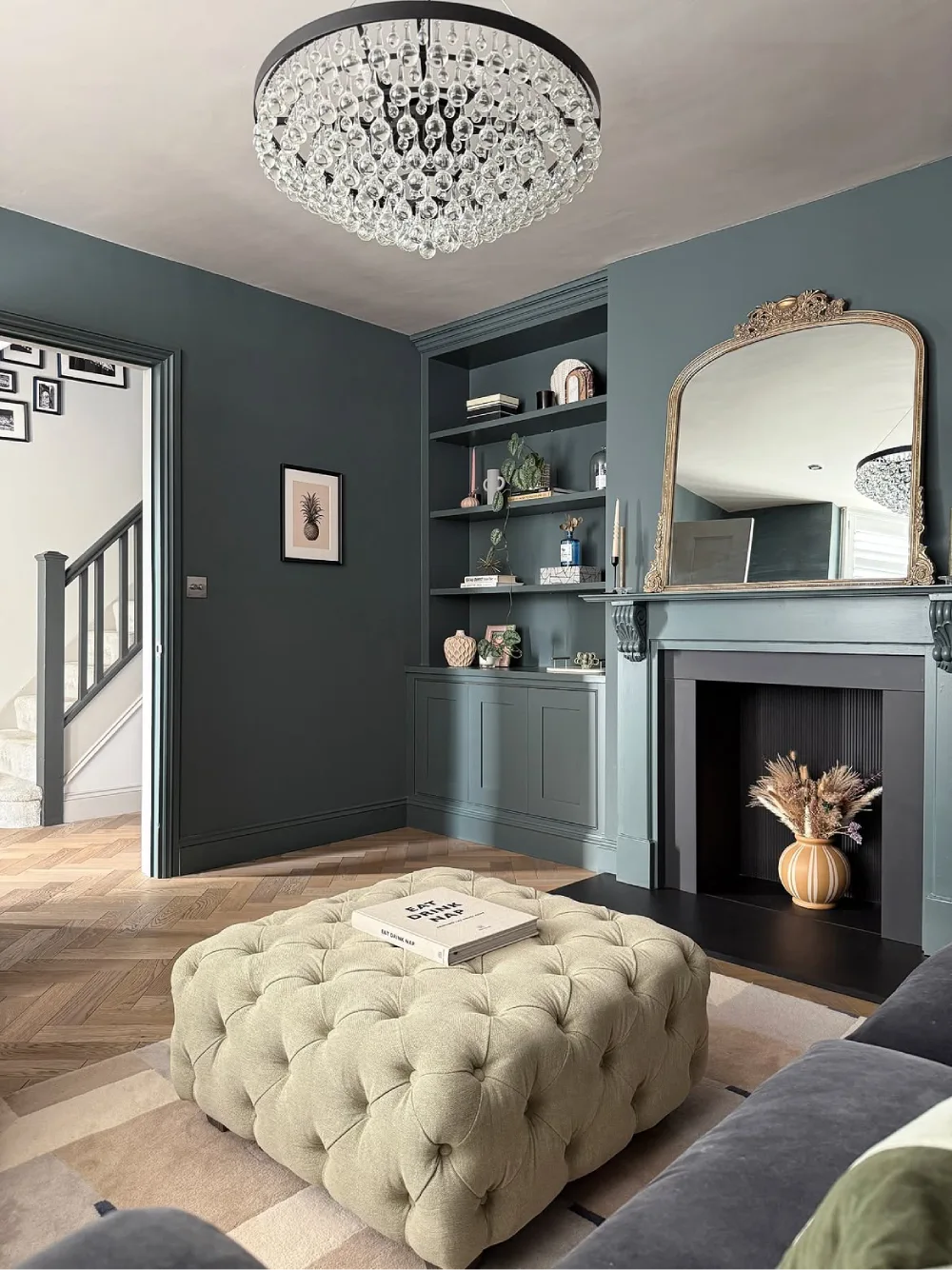 Elegant living room with muted blue-green walls, matching built-in shelves, a cream tufted ottoman, and a decorative gold mirror