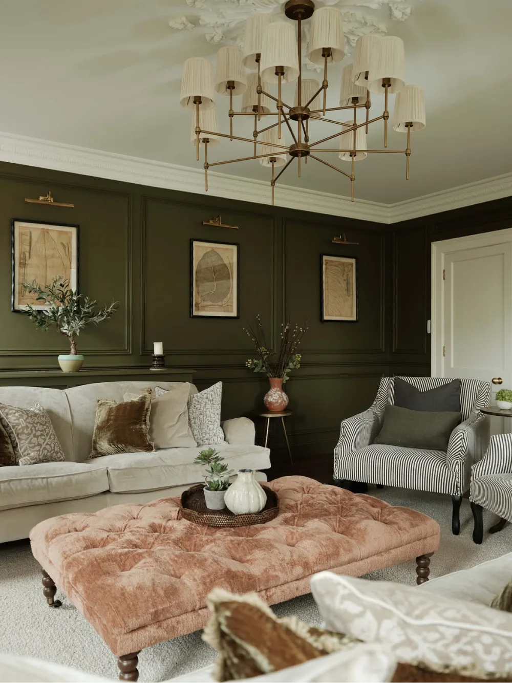 Elegant living room with olive green paneled walls, cream sofas, striped accent chairs, and a blush pink tufted ottoman