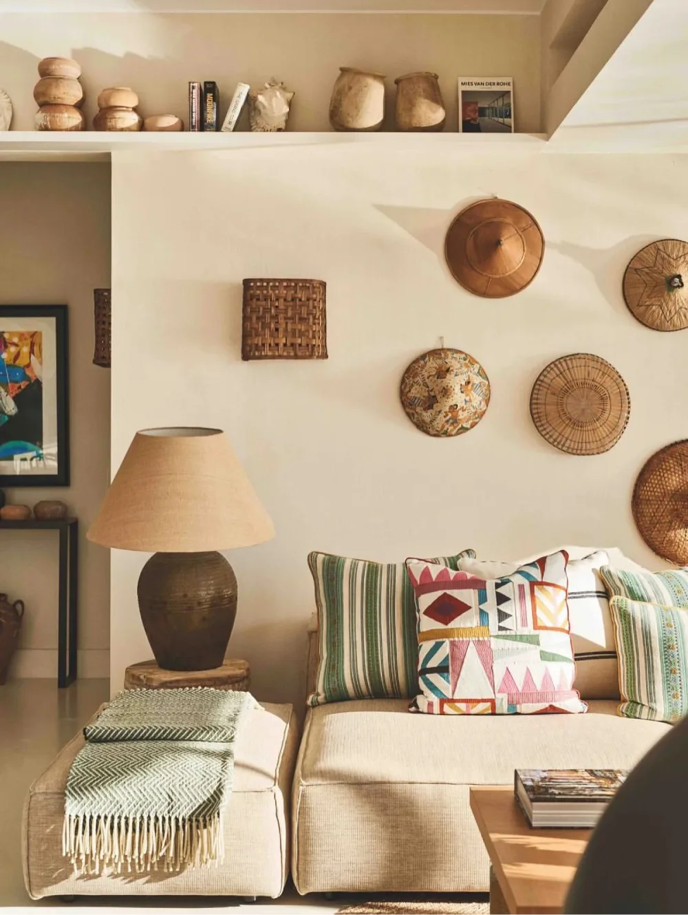 Boho living room with a beige sofa, colorful geometric pillows, woven wall baskets, a ceramic table lamp, and natural wood accents.