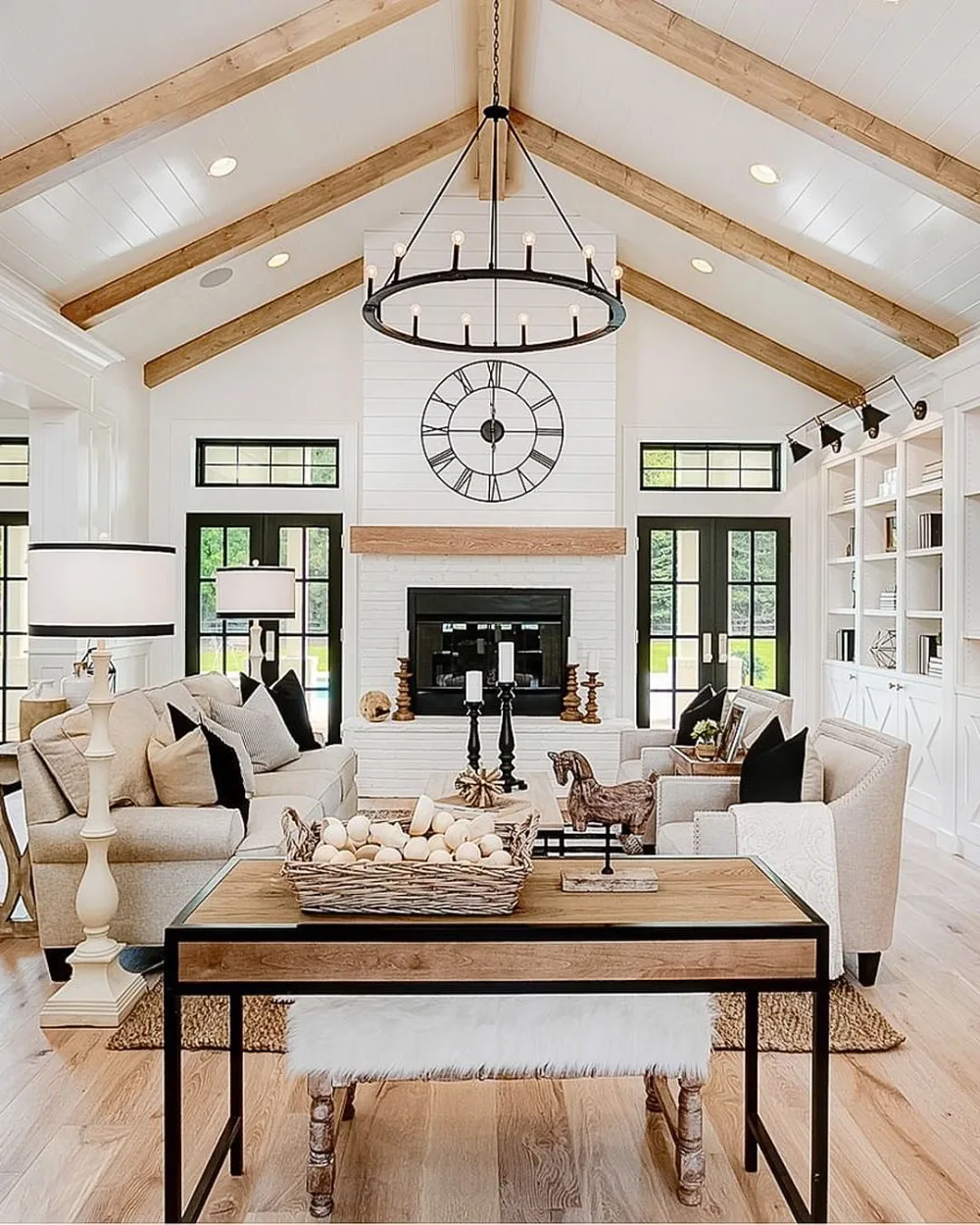 Farmhouse living room with vaulted white ceilings, exposed wooden beams, a black circular chandelier, white sofas, and a central fireplace