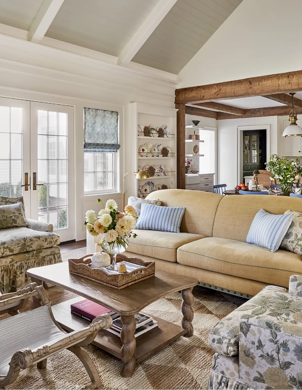 Farmhouse living room with a beige sofa, floral accent chairs, a rustic wooden coffee table, and exposed ceiling beams