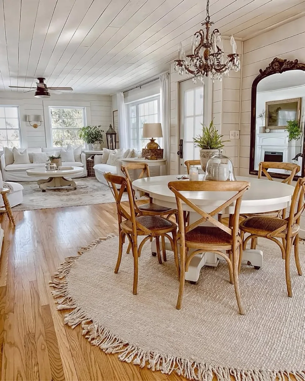 Open-concept farmhouse living and dining room with white shiplap walls, a round dining table, wooden chairs, and neutral area rugs
