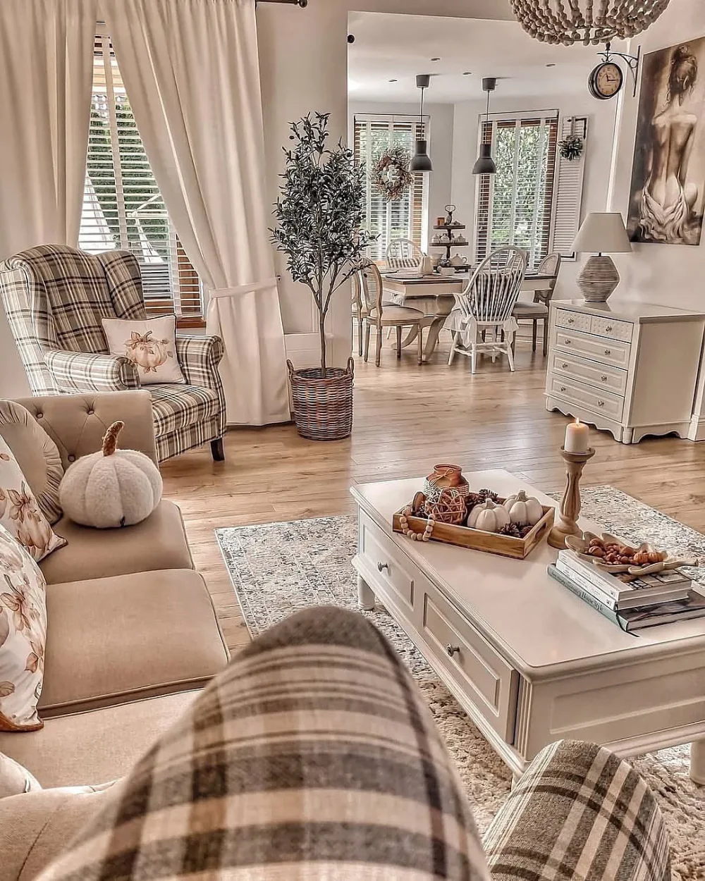 Neutral farmhouse living room with a beige sofa, plaid accent chair, cream curtains, white coffee table, and seasonal pumpkin decorations