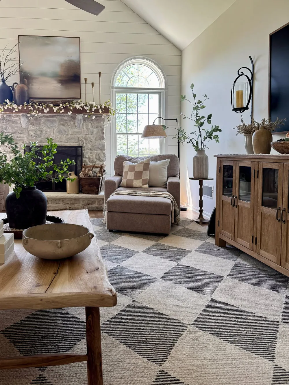 Rustic farmhouse living room with a gray and cream checkered rug, stone fireplace, wooden cabinet, and neutral upholstered armchair