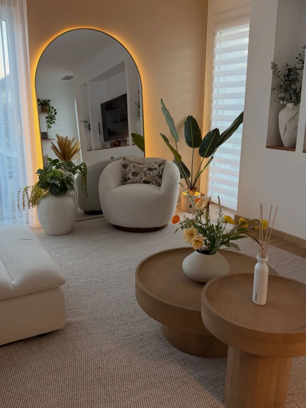 Small living room with a neutral sofa, round coffee tables, plants, and an illuminated arched mirror
