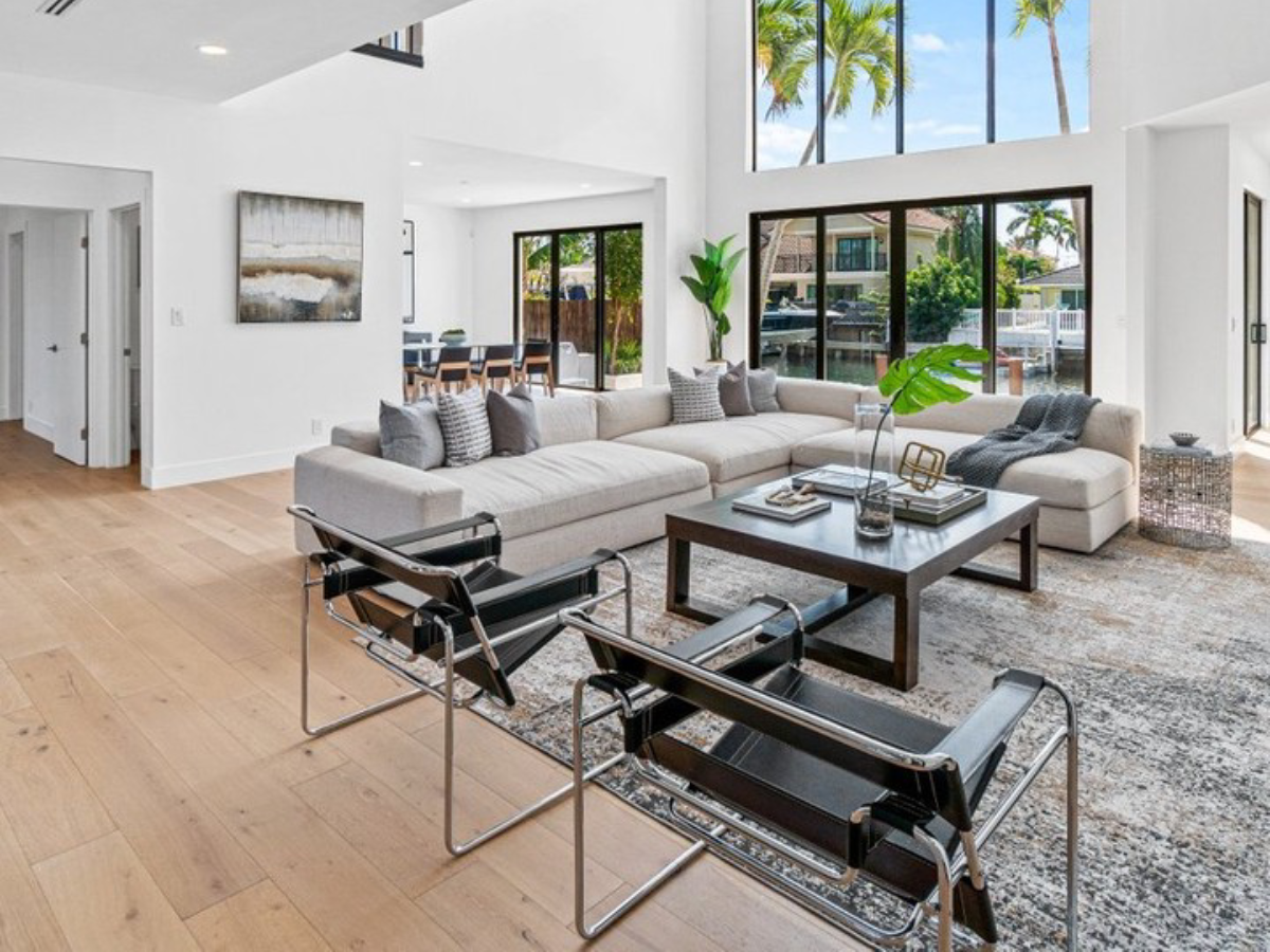 Bright open-concept living room with a large sectional sofa, black leather chairs, wood flooring, and floor-to-ceiling windows overlooking palm trees.