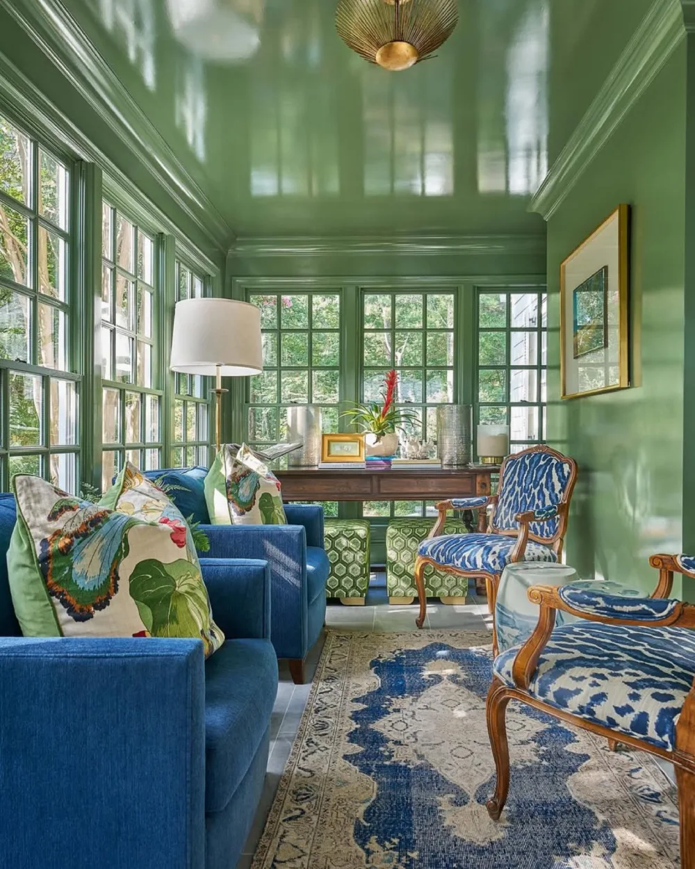 Green sunroom with blue sofas, botanical pillows, patterned chairs, and large windows overlooking trees