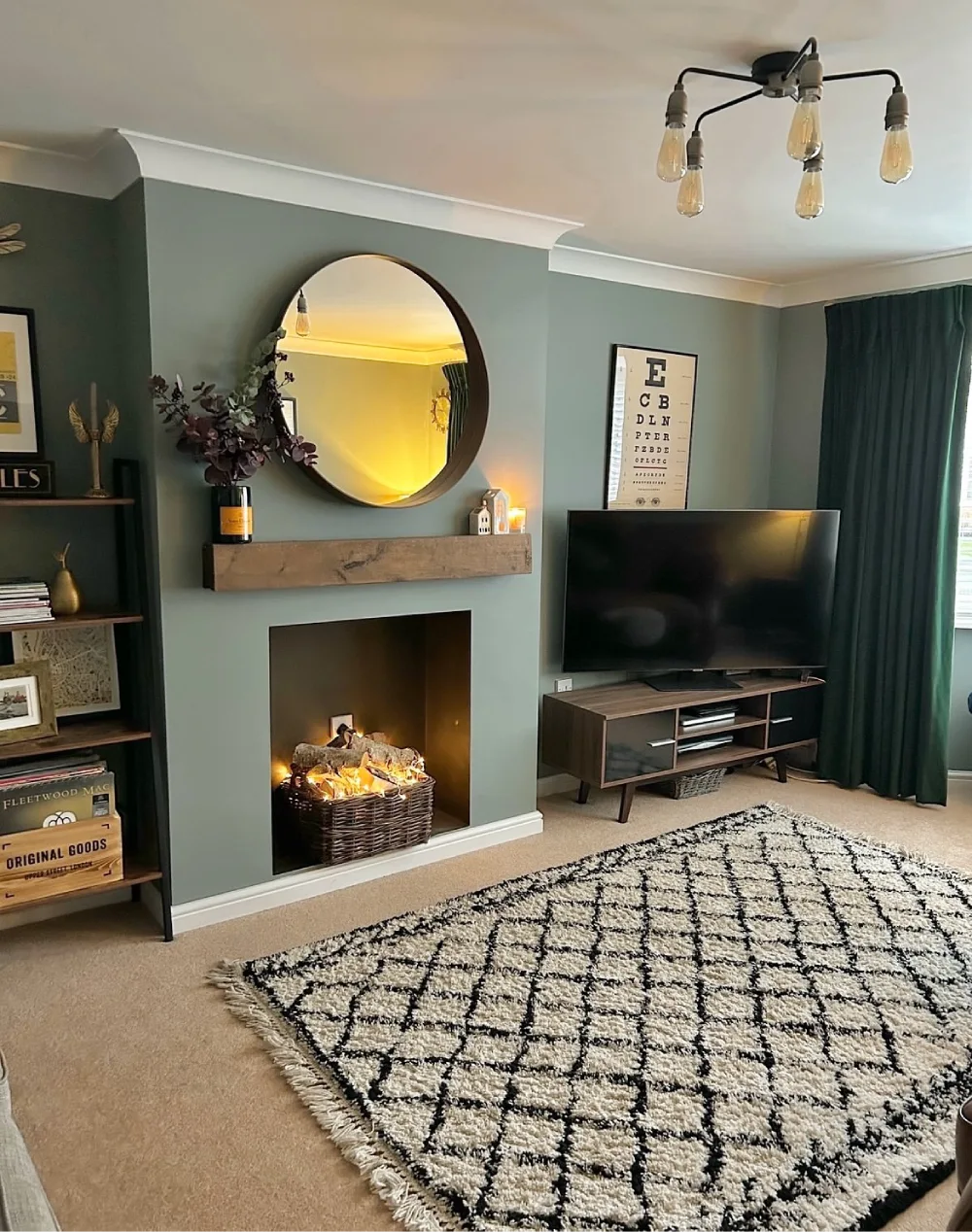 Muted green living room with fireplace, round mirror, dark wood media console, and patterned rug
